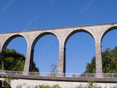 gigantic impressive railway viaduct without train in front of blue sky and green forest with a highway and guardrails by day without people