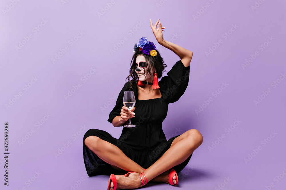 Mischievous Spanish girl with painted face elegantly raised her hand. Woman with crown of flowers drinking champagne sitting in lilac studio Mischievous Spanish girl with painted face elegantly raised her hand. Woman with crown of flowers drinking champagne sitting in lilac studio