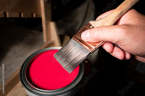 Man's hand prepares to dip paintbrush into one gallon can of red latex paint.