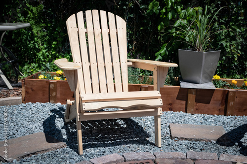 Unpainted wooden Adirondack chair sits in sunshine filled backyard garden courtyard.