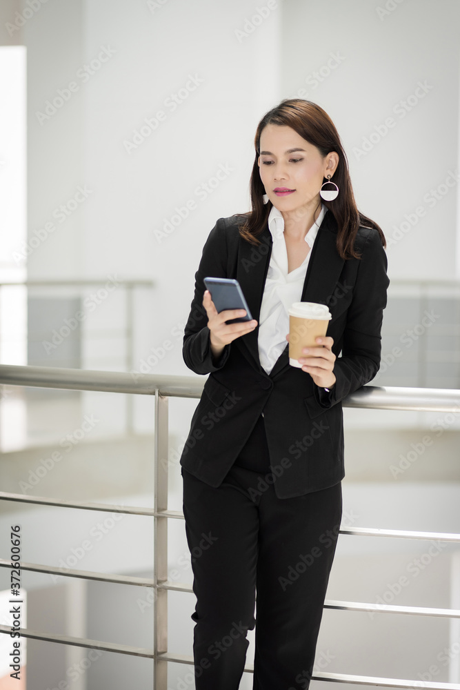 Portrait of a confident businesswoman in business suit holding a cup of coffee while using cellphone during break-time in the business building. Business stock photo. Portrait of a confident businesswoman in business suit holding a cup of coffee while using cellphone during break-time in the business building. Business stock photo.