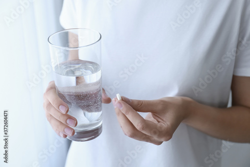 Woman holding glass of water and pill near window at home, closeup