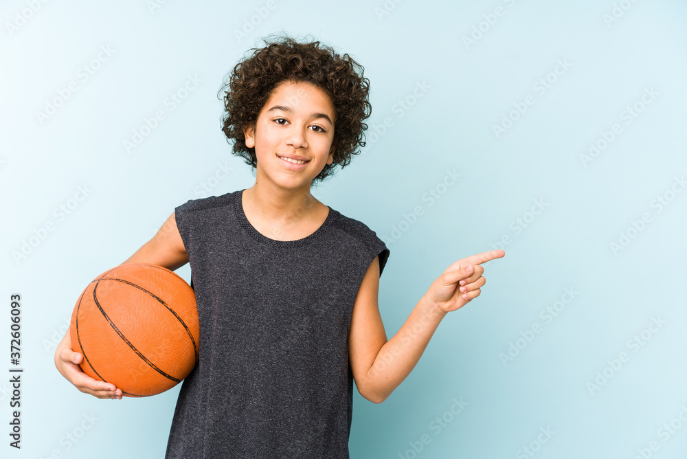 Kid boy playing basketball isolated on blue background smiling and pointing aside, showing something at blank space.