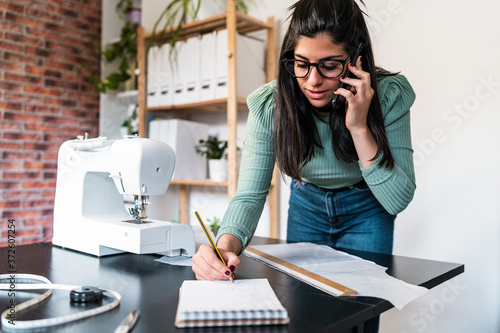 Young ethnic seamstress in eyeglasses taking notes standing leaned forward while using straightedge at table with papers and scissors in loft style studio photo