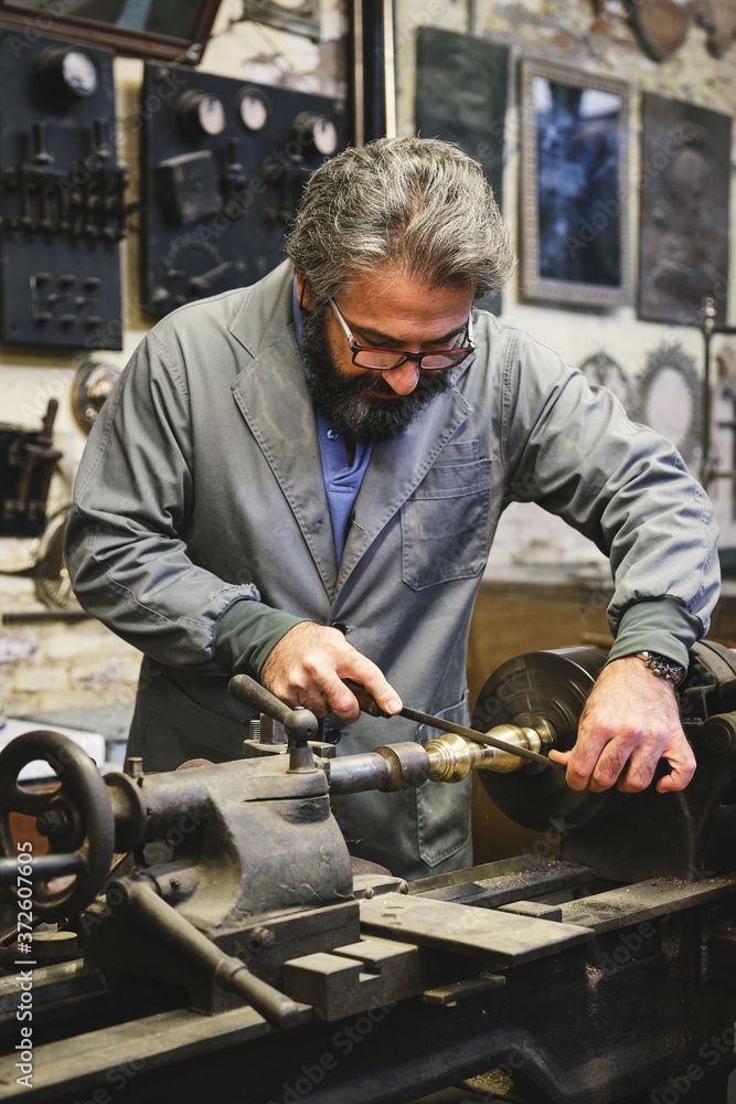 Crop serious mature jeweler in uniform and eyewear sharpening metal stick on professional equipment during work in studio Crop serious mature jeweler in uniform and eyewear sharpening metal stick on professional equipment during work in studio