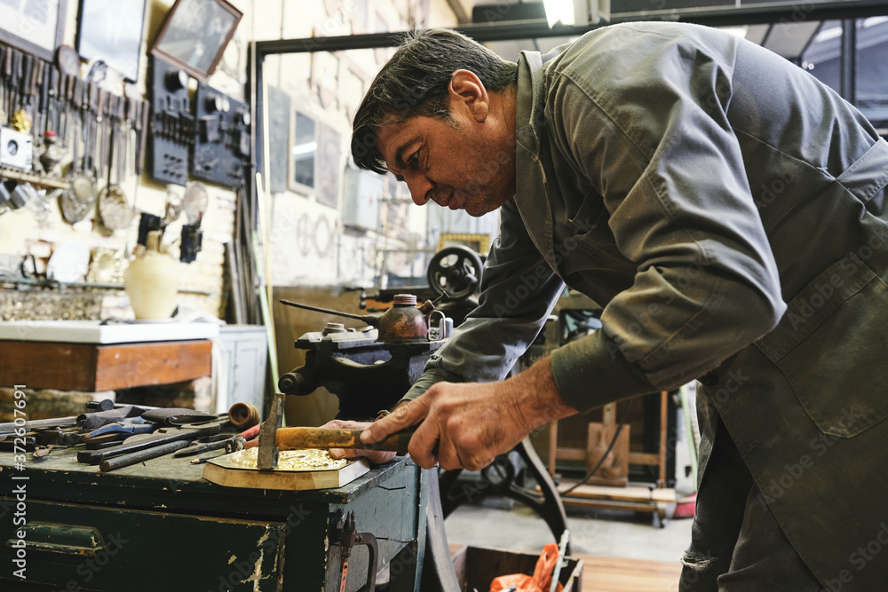 Crop male goldsmith hitting metal detail with hammer during work in workshop Crop male goldsmith hitting metal detail with hammer during work in workshop