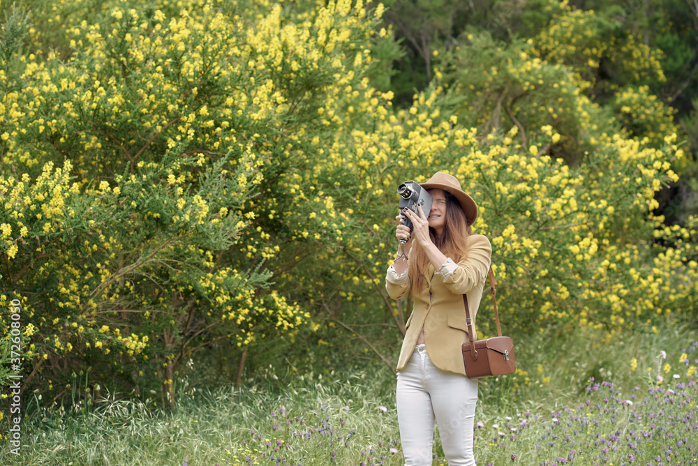 Female photographer in hat with closed eyes filming video with retro video camera while standing near trees with blooming yellow flowers in daylight Female photographer in hat with closed eyes filming video with retro video camera while standing near trees with blooming yellow flowers in daylight