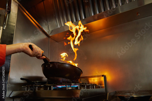 From below cropped unrecognizable chef in uniform holding frying pan while flambeing ingredients in frying pan on slow heat standing in restaurant kitchen in evening and looking at bright fire