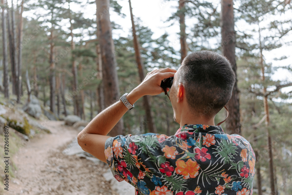 Back view of male tourist in colorful shirt standing on slope of mountain covered with coniferous forest and observing environment with binoculars during hiking in Navacerrada in Spain Back view of male tourist in colorful shirt standing on slope of mountain covered with coniferous forest and observing environment with binoculars during hiking in Navacerrada in Spain