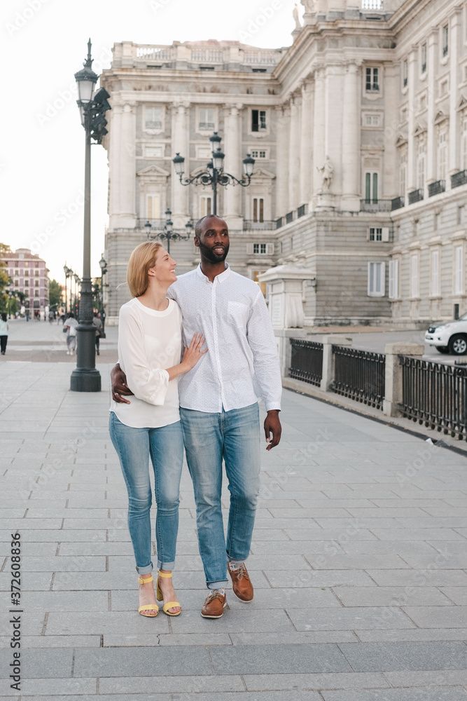 Full body cheerful multiracial couple in jeans and white shirts walking on pavement along street in Madrid while bonding and chatting Full body cheerful multiracial couple in jeans and white shirts walking on pavement along street in Madrid while bonding and chatting