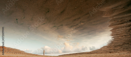 Full body anonymous person in beige casual clothes standing on sandy dunes in dry desert valley reflecting in cloudy sky mirage in desert