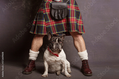 Crop anonymous man in Scottish skirt and leather boots with leather shoulder bag standing with legs apart near cute French Bulldog in red collar