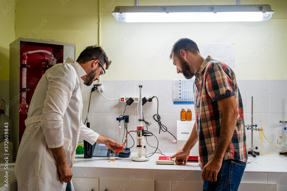 Side view of bearded chemist in eyeglasses standing near coworker in casual clothes in front of wine analysis equipment and flasks in lab Side view of bearded chemist in eyeglasses standing near coworker in casual clothes in front of wine analysis equipment and flasks in lab