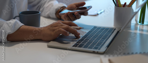 Side view of a man holding smartphone while using laptop on working desk in home office