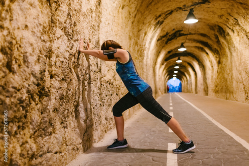 Side view of anonymous fit female athlete in sports clothes and armband standing leaned against rough wall while stretching legs during warm up in tunnel with artificial light