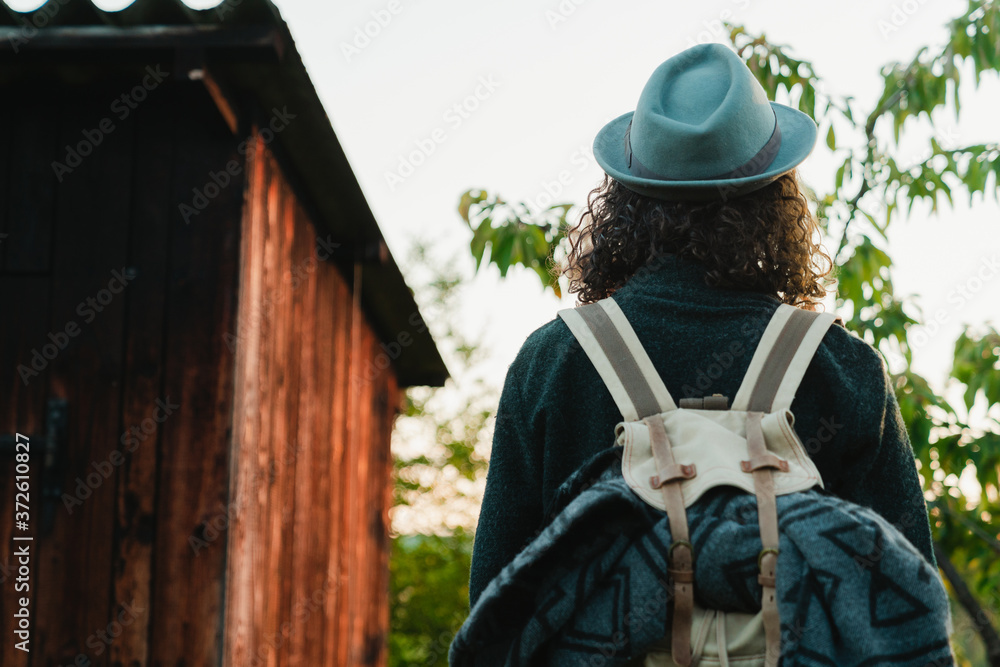 From behind view of anonymous man hipster with long hair and backpack standing in countryside next to a wooden cabin during summer vacation From behind view of anonymous man hipster with long hair and backpack standing in countryside next to a wooden cabin during summer vacation