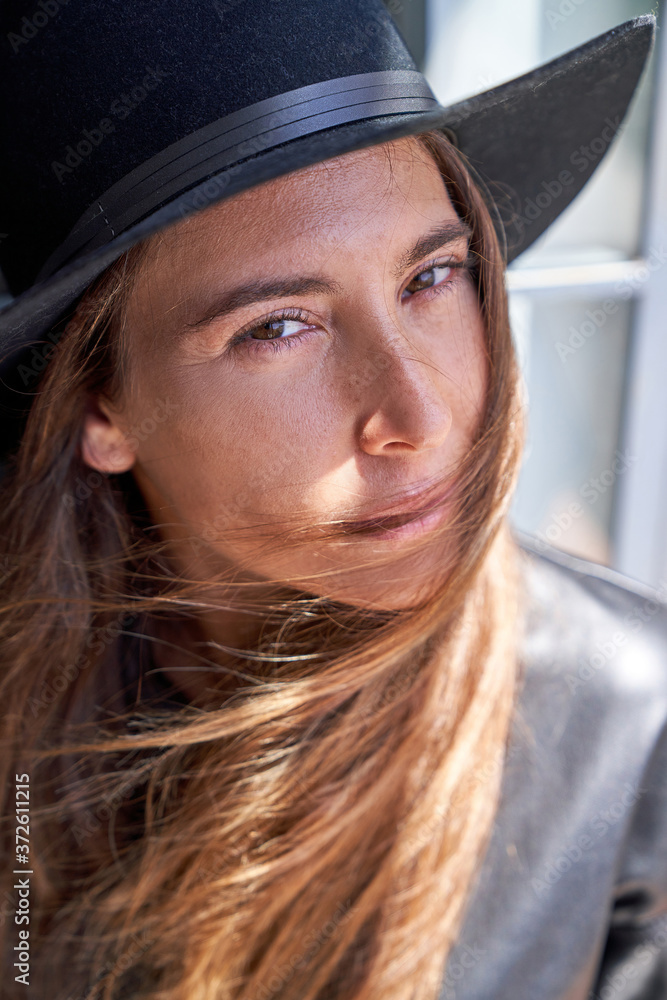 Stylish young long haired female in trendy hat and leather jacket looking away while standing near wall on sunny street Stylish young long haired female in trendy hat and leather jacket looking away while standing near wall on sunny street
