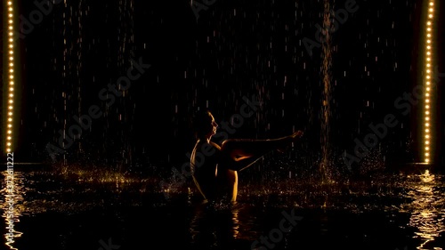 Ubhaya Padangusthasana performed by a flexible woman. The rain pours on the half-naked body of the athlete. Yellow soft light in a dark studio with a black background. Slow motion. photo