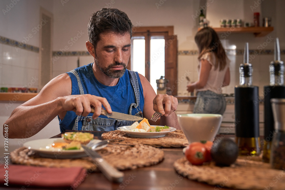 Calm male sitting at table in kitchen and enjoying tasty breakfast on blurred background of female Calm male sitting at table in kitchen and enjoying tasty breakfast on blurred background of female