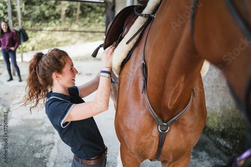 Side view of female equestrian putting saddle on chestnut horse while preparing for dressage photo