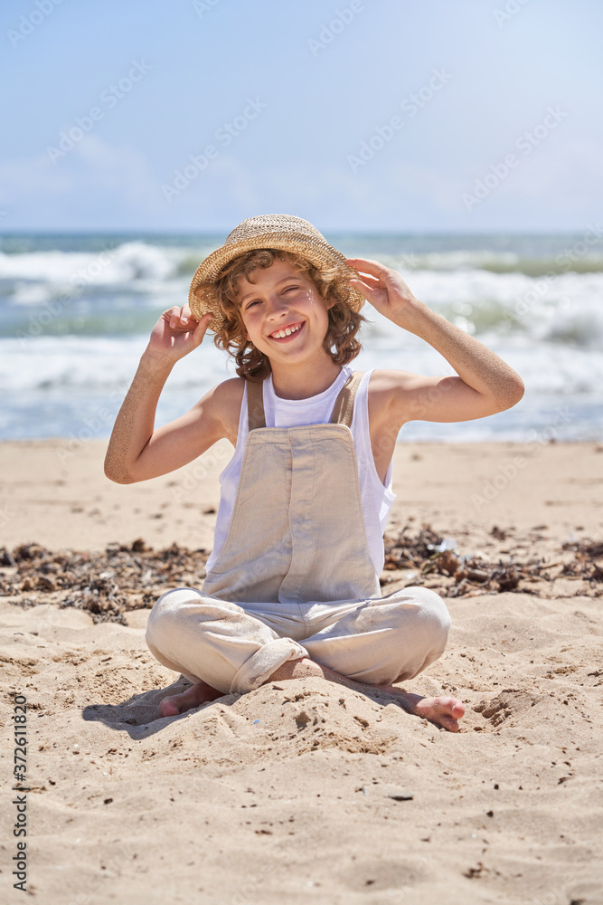 Full body of happy little boy sitting with legs crossed on sandy beach near waving sea Full body of happy little boy sitting with legs crossed on sandy beach near waving sea