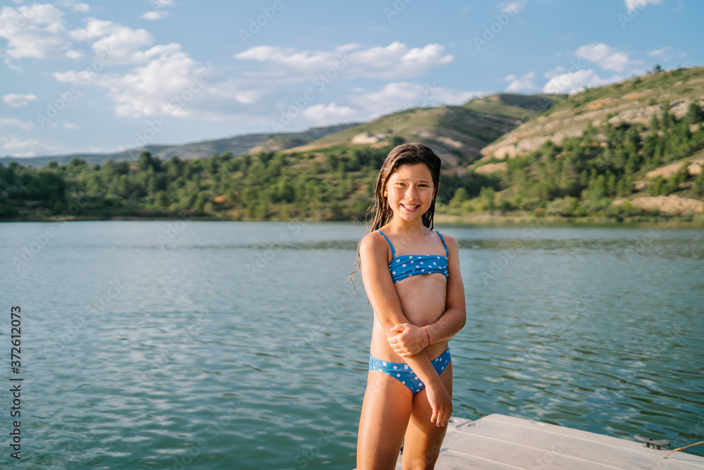 Happy little girl in bikini standing looking at camera in wooden pier near pond during summer holiday Happy little girl in bikini standing looking at camera in wooden pier near pond during summer holiday