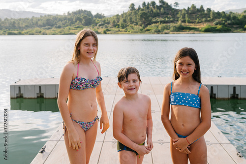 Smiling brother and sisters in swimwear standing on quay near lake and looking at camera while enjoying summer vacation photo