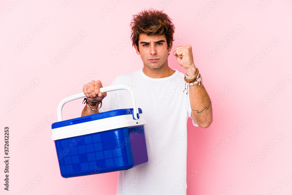 Young caucasian man holding a portable fridge showing fist to camera, aggressive facial expression. Young caucasian man holding a portable fridge showing fist to camera, aggressive facial expression.