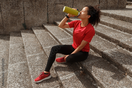 Side view of fit ethnic female athlete in sports clothes and smart watch drinking water from thermos while resting with closed eyes on staircase after training in sunlight