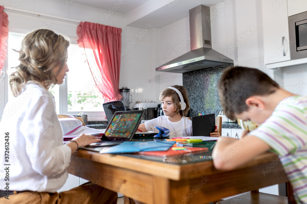 Concentrated mother in formal wear sitting leaned on hand while typing on netbook near unrecognizable son in headset and boy doing homework in light flat Concentrated mother in formal wear sitting leaned on hand while typing on netbook near unrecognizable son in headset and boy doing homework in light flat