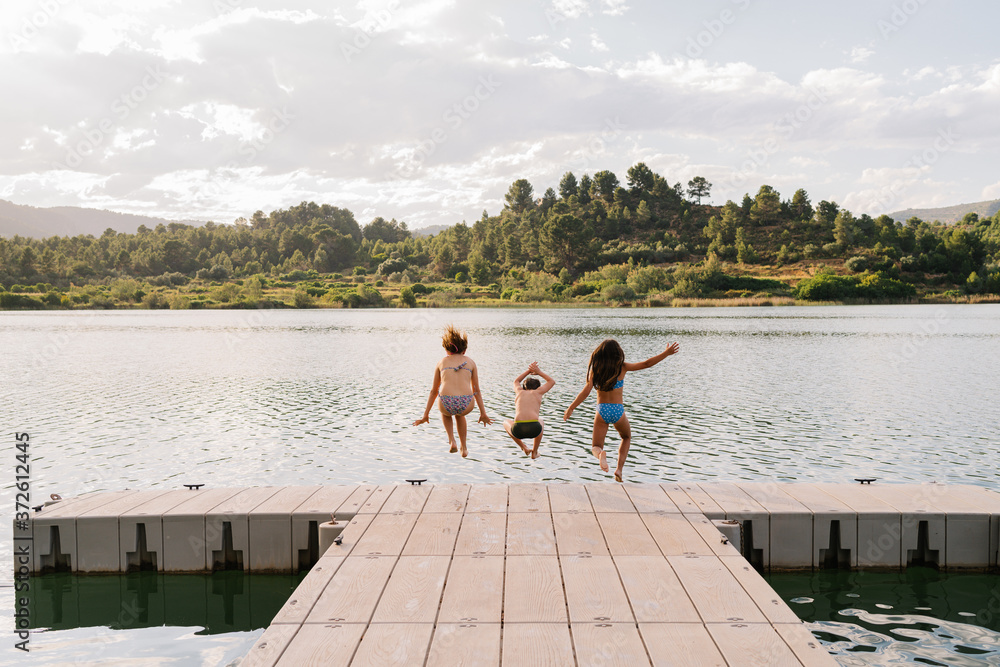Back view of children in swimwear in moment of jumping in pond from wooden quay during weekend in summer Back view of children in swimwear in moment of jumping in pond from wooden quay during weekend in summer