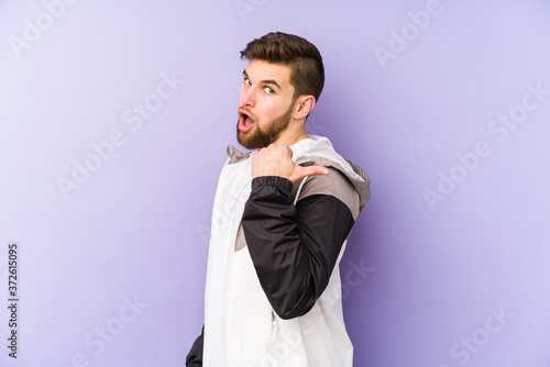 Young man isolated on purple background points with thumb finger away, laughing and carefree.