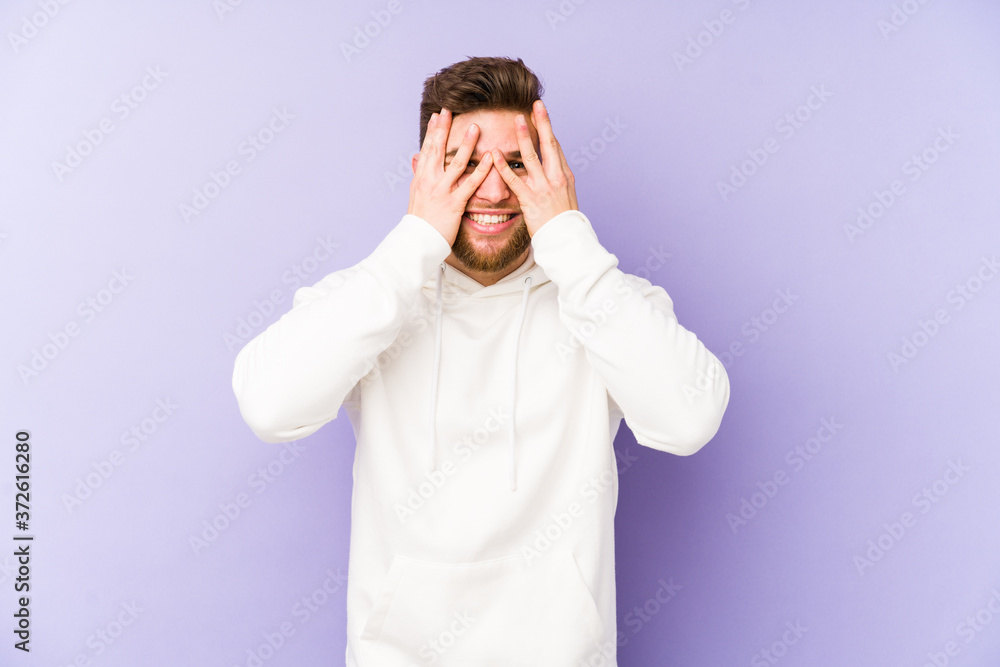 Young caucasian man isolated on purple background blink at the camera through fingers, embarrassed covering face. Young caucasian man isolated on purple background blink at the camera through fingers, embarrassed covering face.
