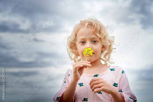 Portrait of pretty young small blonde girl with curly hair in pink dress in field with grass and flowers in summer day and cloudy sky background