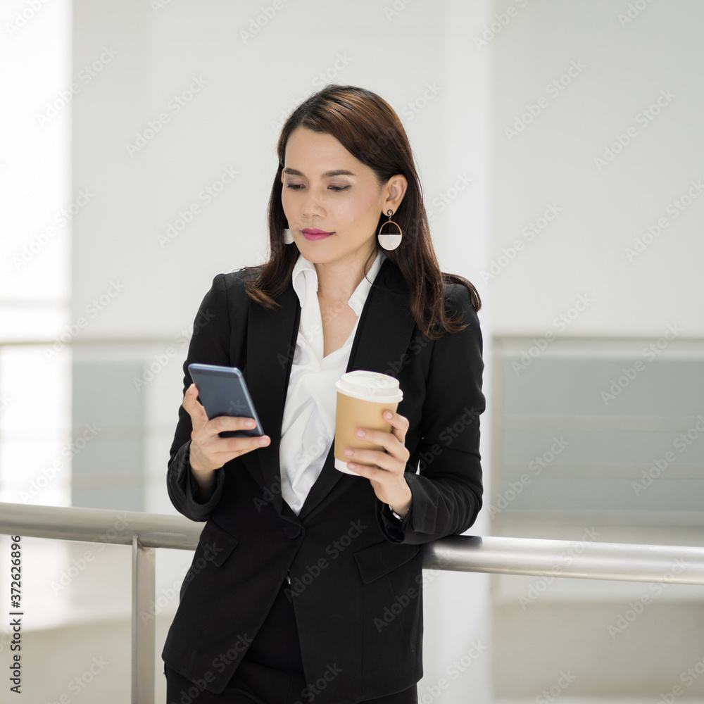 Portrait of a confident businesswoman in business suit holding a cup of coffee while using cellphone during break-time in the business building. Business stock photo. Portrait of a confident businesswoman in business suit holding a cup of coffee while using cellphone during break-time in the business building. Business stock photo.