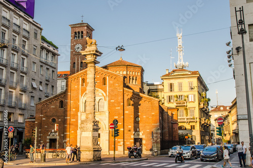 Milan, Italy, September 8, 2018: Basilica di San Babila church with clock tower and column with lion on top between modern buildings on Piazza Babila square in historical centre of Milano city
