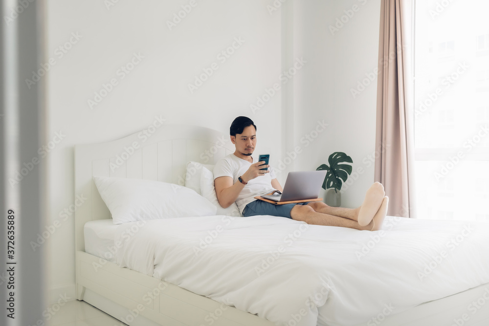 Asian man lying on bed work on his laptop in cozy white bedroom. Asian man lying on bed work on his laptop in cozy white bedroom.