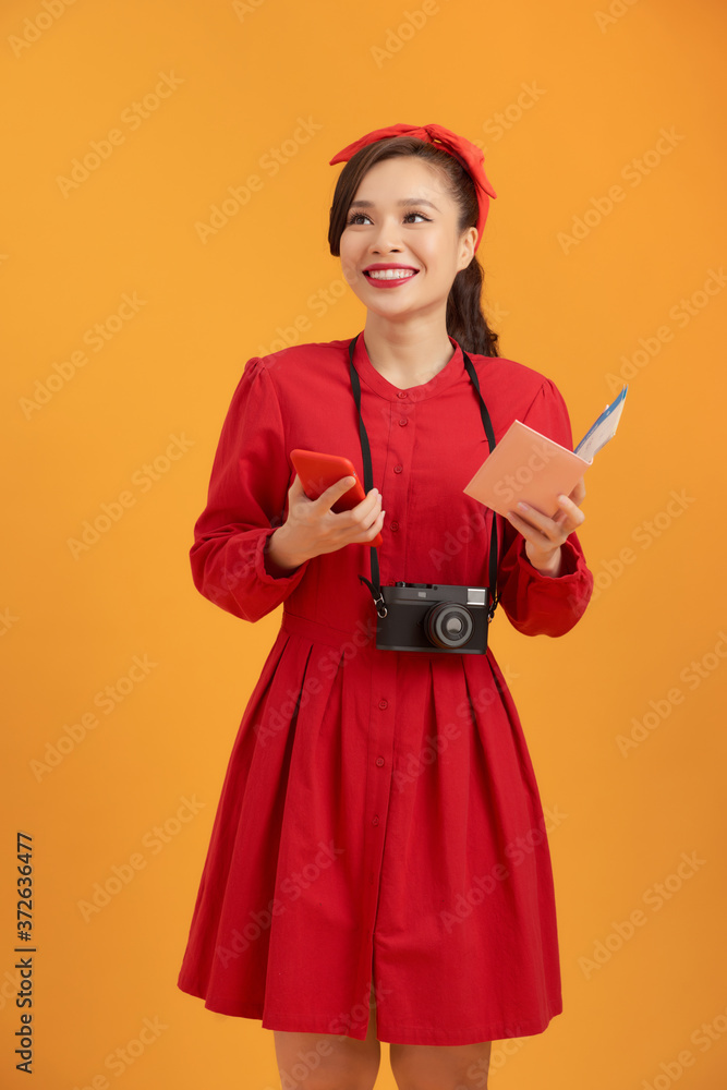 Happy young Asian woman tourist holding phone, passpoer, camera and standing over orange background. Happy young Asian woman tourist holding phone, passpoer, camera and standing over orange background.