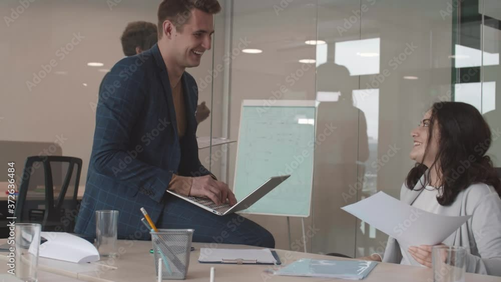 Medium shot of mixed-race businesswoman and Caucasian businessman sitting at desk in modern office and communicating while their two colleagues walking and talking on background Medium shot of mixed-race businesswoman and Caucasian businessman sitting at desk in modern office and communicating while their two colleagues walking and talking on background