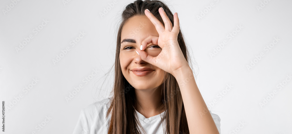 Fototapeta premium Close up portrait of carefree modern girl with long chestnut hairs, looking at the camera through fingers in OKAY gesture. Face expressions, emotions, and body language