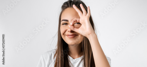 Close up portrait of carefree modern girl with long chestnut hairs, looking at the camera through fingers in OKAY gesture. Face expressions, emotions, and body language