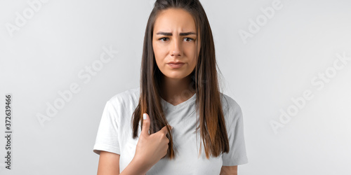 Me. Portrait of indignant girl with long chestnut hair startled by offensive words, points at herself with for finger, opens mouth in surprisement, has dissatisfied expression, wears casual clothes photo