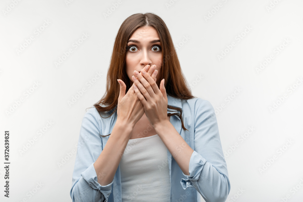 Shocked caucasian girl with long chestnut hair, dressed in casual clothes, covering mouth with hands on white background. Keep silence concept.