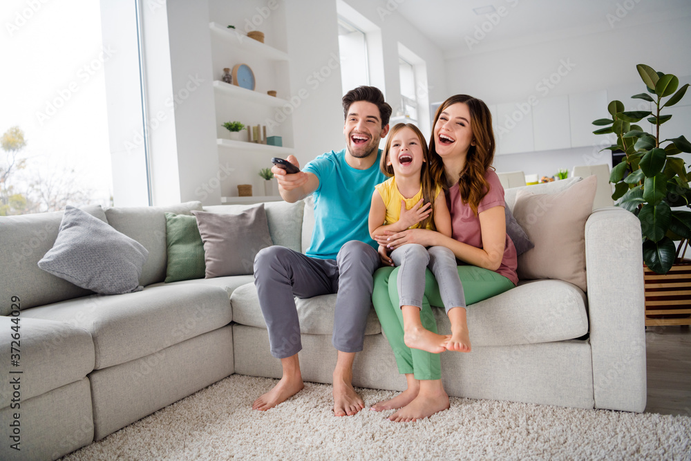Full size photo of positive three people mom dad preteen little kid girl sit comfort couch rest watch comedy cartoon remote control switch joy laugh in house indoors