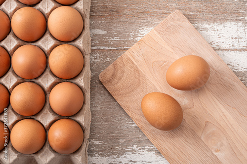 Two eggs on cutting board and many fresh raw brown chicken eggs in carton box on wooden rustic white table background. Top view.