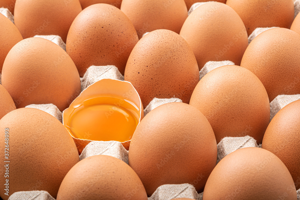 Many fresh raw brown chicken eggs in carton. Beautyful brown chicken egg is half broken with yolk among other eggs, top view.