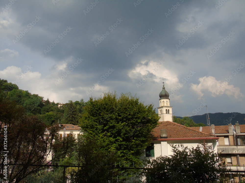 Tarzo, Italy - 08/15/2020: Beautiful photography of a little village from north Italy in summer days. View to the old church, mountains and some houses with grey and blue sky in background.