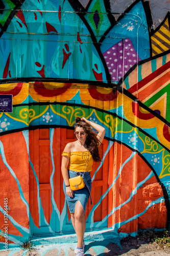 Attractive brunette girl wearing a blue skirt and an orange top posing on a summer day with a colorful wall in the background. Yellow purse, stylish influencer travelling and modelling