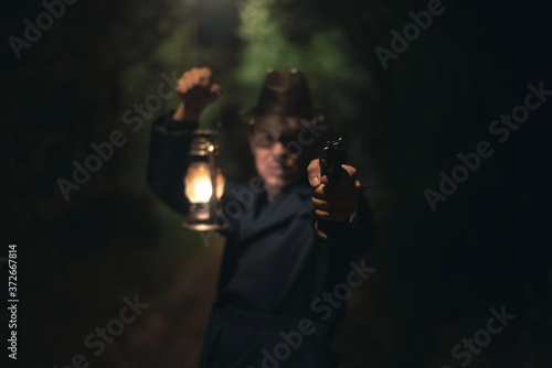 Detective agent in a hat and coat is holding in hand a kerosene lamp and is aiming by a gun into a dark.