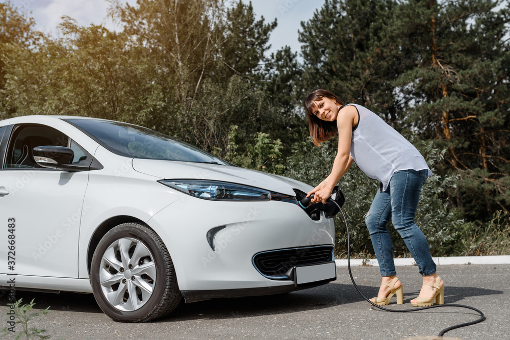 Young beautiful woman is charging her electric car at the charging station situated in the forest. Young beautiful woman is charging her electric car at the charging station situated in the forest.